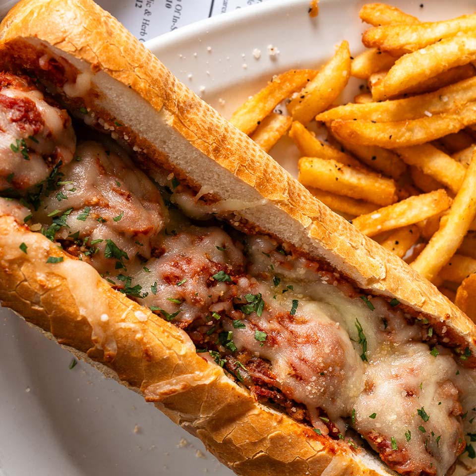 Italian meatball sub with melted cheese, herbs, and seasoned fries served on a white plate.