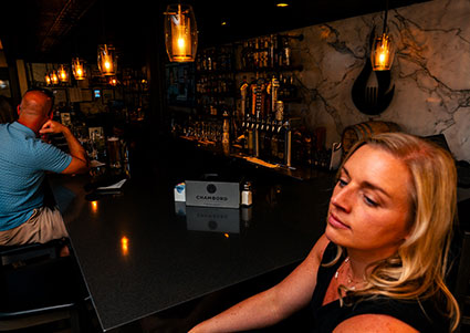 A woman sits at DeBlaze at 131′s dimly lit bar; bottles gleam behind the steakhouse counter.