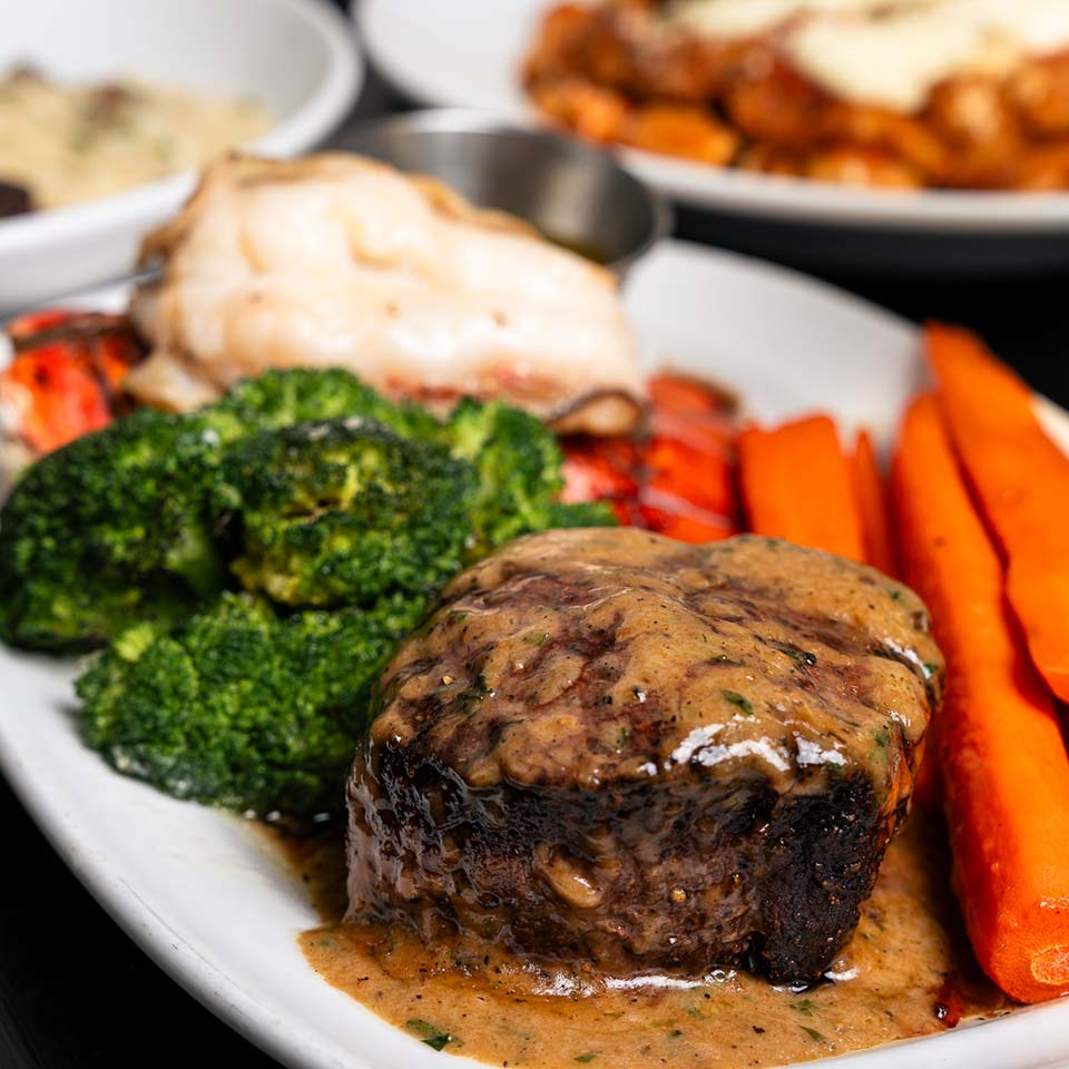 Steakhouse delight: steak with sauce, carrots, broccoli, lobster on white plate, dishes behind.