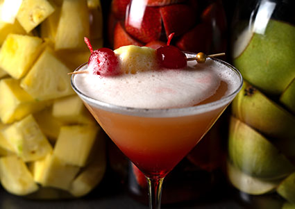 A Bar classic: martini glass with pink frothy drink, cherries, pineapple, and fruits behind.