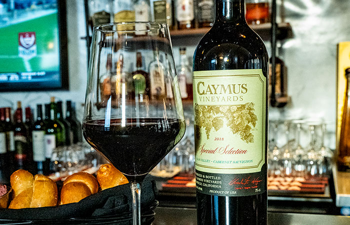 A bottle of Caymus wine and a glass sit on DeBlaze at 131′s bar beside bread rolls.