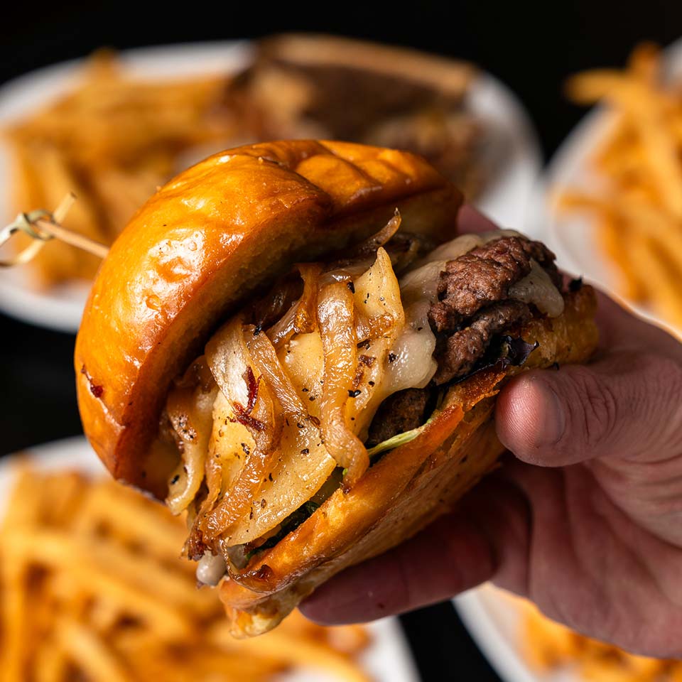 Hand holding a cheeseburger with caramelized onions; Bar plates of fries in the background.