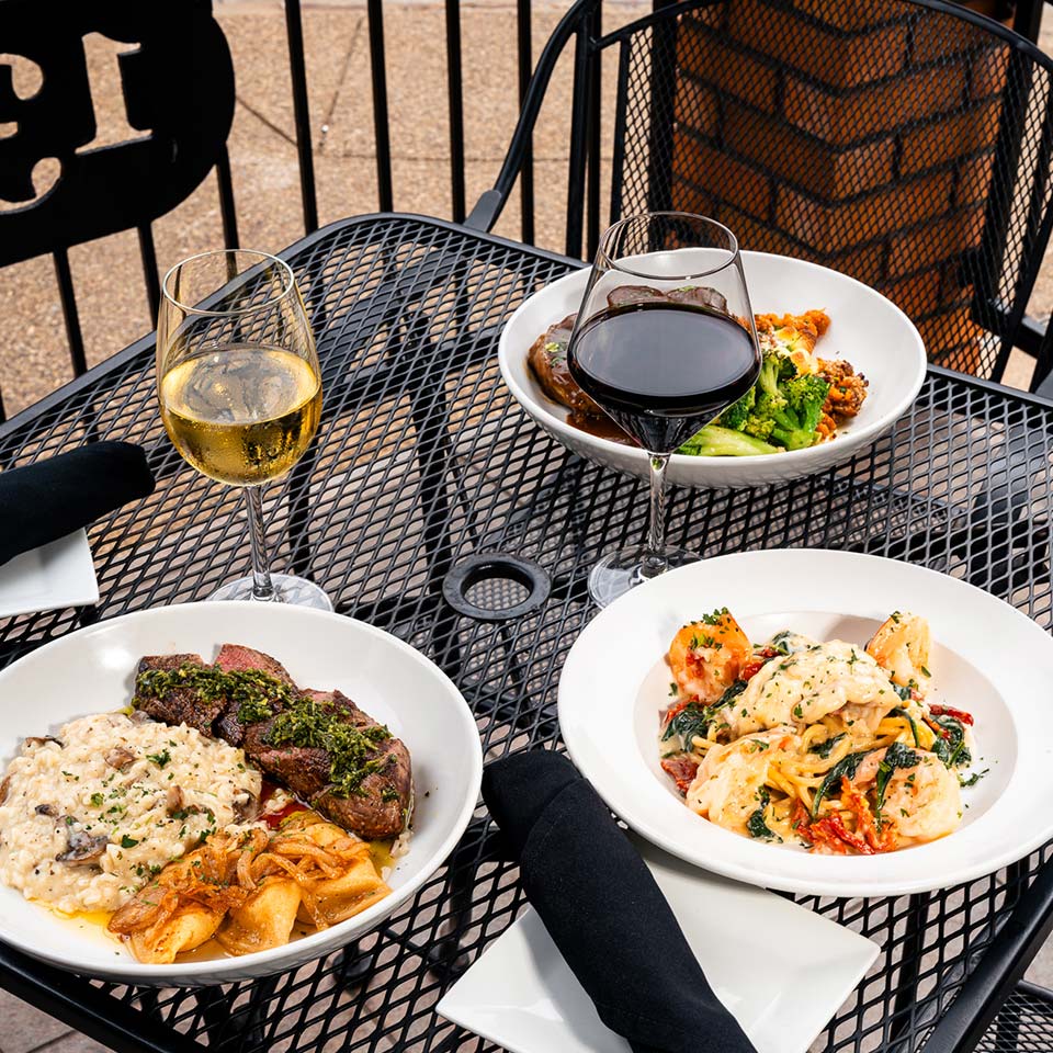 Three plated meals, wine glasses, and black napkins set on a Carnegie Restaurant patio table.