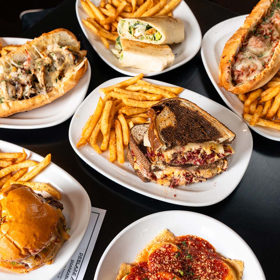 Plates with sandwiches, fries, a wrap, and Italian pasta with red sauce on a black table.