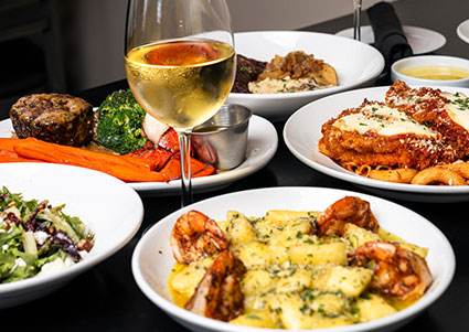 A table at DeBlaze at 131 with shrimp pasta, salad, cutlet, veggies, and white wine.