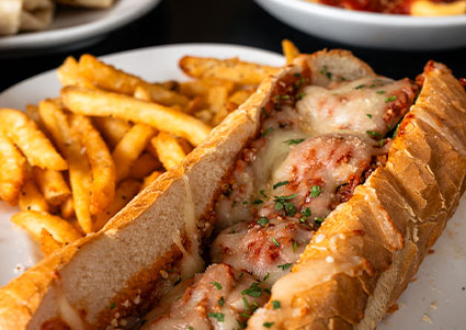 Italian meatball sub with melted cheese, served with seasoned fries on a white plate.